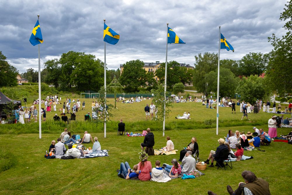 Picknick i gräset framför midsommarstången, med svenska flaggor i förgrunden.